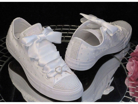 White pearl wedding Converse trainers with satin ribbons and heart diamante sliders on reflective surface.